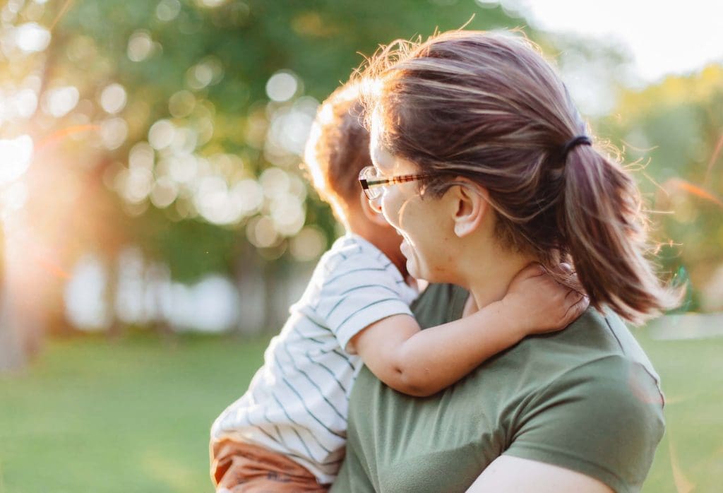 A mother and her young toddler son smiling together at a local park on a sunny summer day.