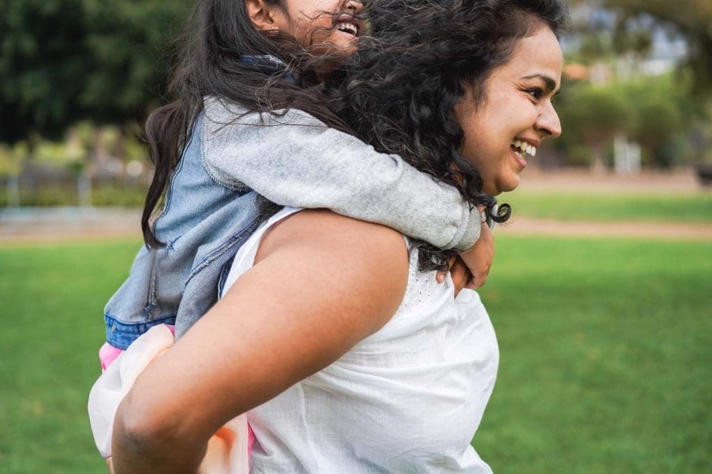 A smiling mother giving her young child a piggyback ride in a sunny park.