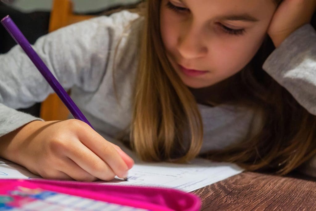A young girl sitting at a wooden table focusing intently on doing her homework.